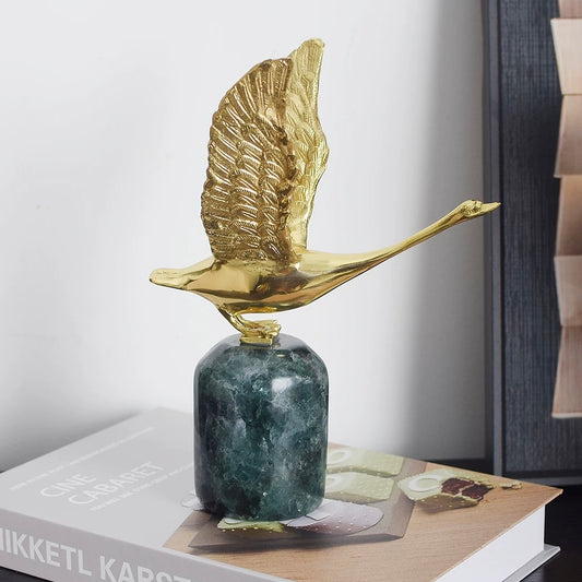 A majestic brass swan sculpture in mid-flight, perched on a deep green fluorite base, styled on a coffee table book.