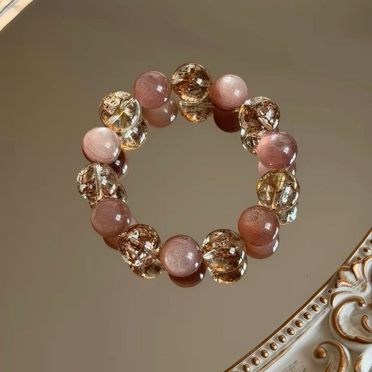 A high-definition product view of SeekEven's natural gold rutilated quartz and pink lepidolite bracelet resting on a pristine mirror, showing rich golden inclusions and smooth lavender-pink crystals