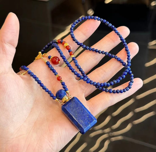 A model's hand holding a lapis lazuli aromatherapy bottle necklace with red agate beads.